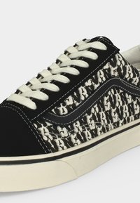 Ασπρόμαυρο χαμηλό αθλητικό παπούτσι με κρεμ κορδόνια, πλευρές από υφασμάτινο ύφασμα με σχέδιο, λεπτομέρεια μαύρης ρίγας και λαστιχένια σόλα εκρού.