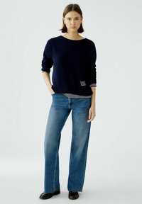 Dunkelblauer Pullover mit langen Ärmeln und rundem Halsausschnitt, über einem gestreiften Hemd getragen. Kombiniert mit weit geschnittenen blauen Jeans und schwarzen Schuhen.