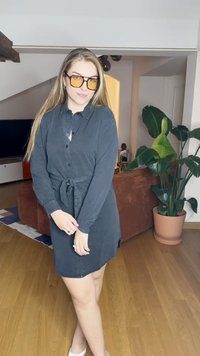 Abito camicia nero con bottoni, vita legata, maniche lunghe e una leggera lucentezza. Indossato con occhiali di grandi dimensioni con tonalità gialle in un ambiente domestico.