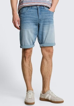 Mann trägt aufgekrempelte hellblaue Jeansshorts, ein gestreiftes T-Shirt und beige Sneaker mit braunen Sohlen und steht vor einem schlichten Hintergrund.