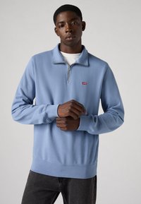 Helles blaues Sweatshirt mit Reißverschluss aus weichem Stoff; ausgestattet mit einem hohen Kragen, langen Ärmeln und einem kleinen roten Levi's-Logo auf der Brust.