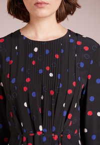 Närbild av en kvinna som bär en svart klänning med vertikala plisseringar och röda, blå och vita prickar på tyget.