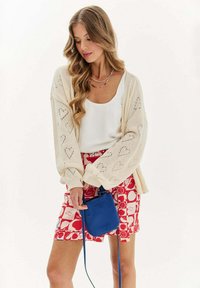 Žena s vlnitými vlasmi, oblečená v bielom tielku, smotanovom pletenom kardigáne s motívom srdca a červených šortkách s vzorom, drží malú modrú crossbody tašku.