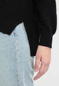 Musta neulottu villapaita, jossa on kohokuvioitu kuvio ja resorireunukset, yhdistetty vaaleansinisiin farkkuihin. Helman sivussa näkyy halkio.