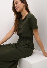 Olivgrünes Kleid mit kurzen Ärmeln, elastischem Taillenbund und fließendem Stoff. Modell sitzt auf einer weißen Fläche, entspanntes Auftreten.