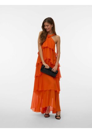 Frau in einem mehrlagigen orangen Maxikleid mit Neckholder, die eine schwarze Clutch hält und schwarze Sandalen mit Absatz trägt, lächelt mit langen, welligen Haaren.