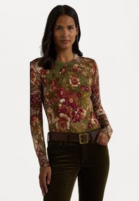 Μακρυμάνικο floral μπλούζα με πράσινο φόντο και κόκκινα και κρεμ λουλούδια, συνδυασμένη με σκούρα πράσινα κοτλέ παντελόνια και καφέ ζώνη.