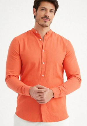 Homme avec une légère barbe portant une chemise orange à manches longues boutonnée, debout devant un fond blanc uni.
