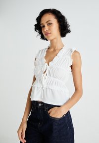 White, ruffled blouse with a v-neck, featuring a drawstring front and layered design. Worn with dark denim jeans.
