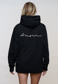 Black hoodie featuring a large embroidered "Amour" in white cursive on the back. Designed with a spacious hood and long sleeves.