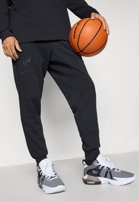 Black Nike sweatpants feature a raised Chicago Bulls logo and tapered design. Paired with black and white athletic shoes, holding a basketball.