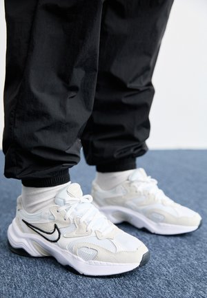 Sneakers atletiche bianchi e grigio chiaro con una tomaia testurizzata, suola spessa e logo swoosh nero. Indossati con pantaloni jogger neri e calzettoni bianchi.