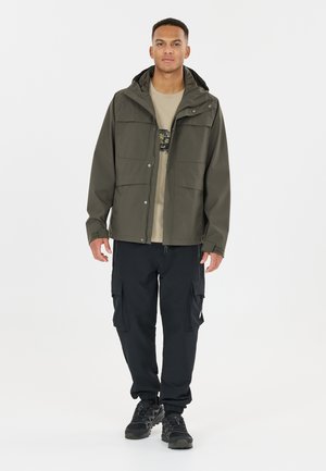 Homme portant une veste à capuche vert olive sur un t-shirt beige à motifs, un pantalon cargo noir et des chaussures de randonnée noires, debout devant un fond blanc uni.