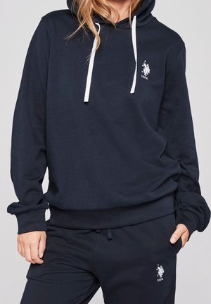 Osoba ubrana w granatową bluzę z kapturem USPA i dopasowane dresy z białymi sznurkami i logo na piersi oraz udzie.