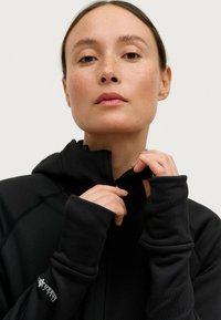 Giacca nera con zip e cappuccio, caratterizzata da una texture liscia e polsini aderenti. I dettagli includono un discreto logo bianco sul maniche.