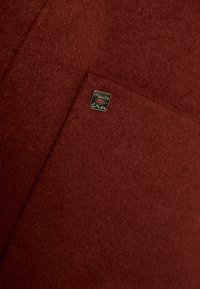 Indumento in feltro rosso con una consistenza liscia, caratterizzato da una piccola etichetta in argento con la scritta "Manila Grace" sulla tasca anteriore.