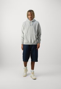 Sweat à capuche gris avec poche avant, shorts en denim et baskets blanches avec accents jaunes ; coupe décontractée, matériau casual et longueur standard.