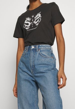 Osoba ubrana w niebieskie jeansy z wysokim stanem oraz czarną koszulkę z białymi wzorami smoka i tekstem na piersi.