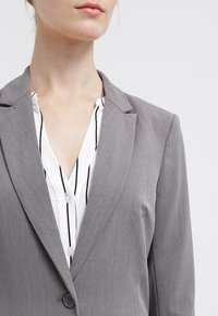 Gray blazer with a notched lapel and single button, worn over a white shirt with black vertical stripes, exhibiting a smooth texture.