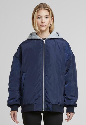 Marineblå bomberjakke med en lys grå hætte, der har en lynlås foran, sidelommer og ribbede manchetter og i bunden.