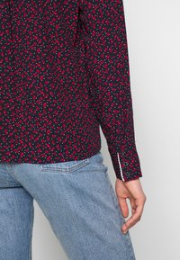 Camicia a maniche lunghe in tessuto navy con un motivo floreale rosso e bianco. Presenta polsini con bottoni e bordi a contrasto, e una vestibilità rilassata.
