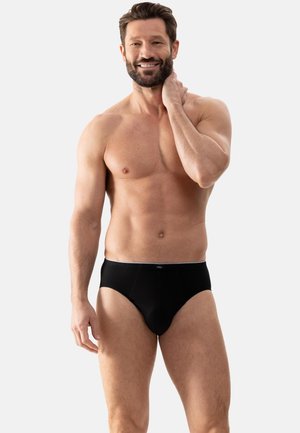 Volwassen man glimlacht, zonder shirt, draagt zwarte slips, staand tegen een effen grijze achtergrond met één hand die zijn nek aanraakt.