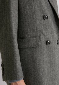 Grijze wollen blazer met een subtiel gestreept patroon, dubbelzijdig ontwerp en grote zwarte knopen. Inclusief een detail van een zijzak.