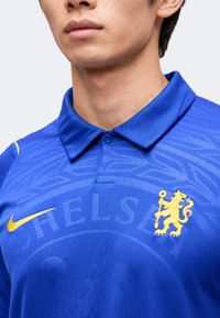 Άνδρας που φοράει μπλε ποδοσφαιρική φανέλα Chelsea της Nike με κίτρινο λιονταράκι στο έμβλημα και διακριτικό μοτίβο λογότυπου της ομάδας στο ύφασμα.