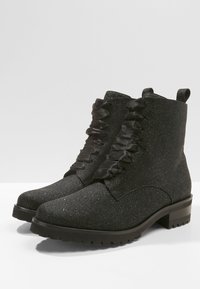 Botines negros con textura brillante, cintas de cinta, punta redondeada y un tacón bajo y resistente. Presenta una lengüeta en la parte posterior para facilitar su uso.