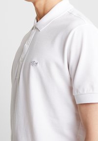 Polo blanc en coton, doté d'un col classique, d'une patte à cinq boutons et d'un petit logo brodé sur le côté gauche de la poitrine.