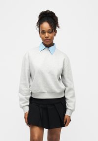 Jeune femme aux cheveux bouclés attachés en chignon, portant un sweat-shirt gris clair, une chemise bleue à col, et une jupe plissée noire, debout devant un fond blanc.