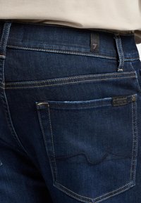 Dunkelblaue Denim-Jeans mit Kontrastnähten, die eine Gesäßtasche mit einem dekorativen Nahtmuster und einem kleinen Markenlabel aufweist.