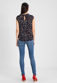 Blusa blu navy con motivo floreale in rosso e bianco, dettaglio a chiave sulla schiena, abbinata a jeans aderenti azzurri e tacchi alti rossi.