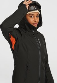 Veste de ski noire avec une finition texturée, présentant une fermeture éclair à l'avant, un accent orange sur la manche, et un col haut avec une capuche ajustable.