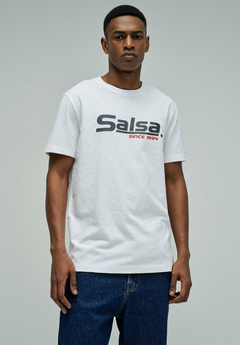 Man draagt een wit T-shirt met de tekst "Salsa sinds 1994" en een blauwe spijkerbroek, staand tegen een effen lichtgrijze achtergrond.