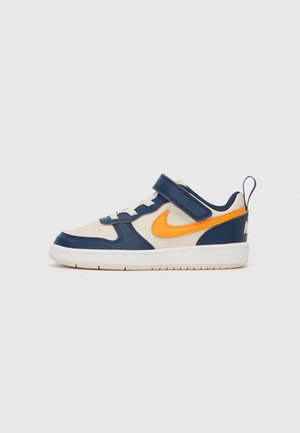 Lage kindersneaker met witte en marineblauwe panelen, oranje Nike-swoosh, witte zool, veters en marineblauwe klittenbandsluiting.