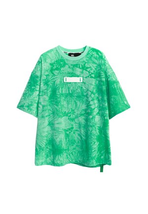 Persona con camiseta verde tie-dye con grandes alas negras y plateadas en la espalda, pantalones beige con diseños de alas azules y verdes, y botas blancas.