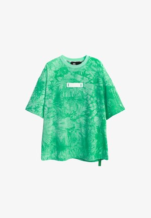 Persona con camiseta verde tie-dye con grandes alas negras y plateadas en la espalda, pantalones beige con diseños de alas azules y verdes, y botas blancas.