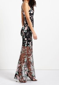 Robe maxi noire avec broderies florales en rose et vert. Caractéristiques d'une silhouette ajustée et d'une jupe transparente, avec des fines bretelles et une fente sur le côté.