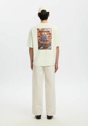 Mężczyzna w kremowych spodniach, koszulce z grafiką retro głośnika i regału z książkami na plecach, kremowej czapce bejsbolowej i ciemnych butach, stojący tyłem.