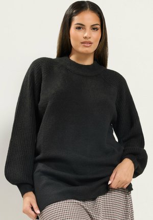 Femme portant un pull en maille noir avec des manches bouffantes et un pantalon à motif pied-de-poule, debout devant un fond clair uni.
