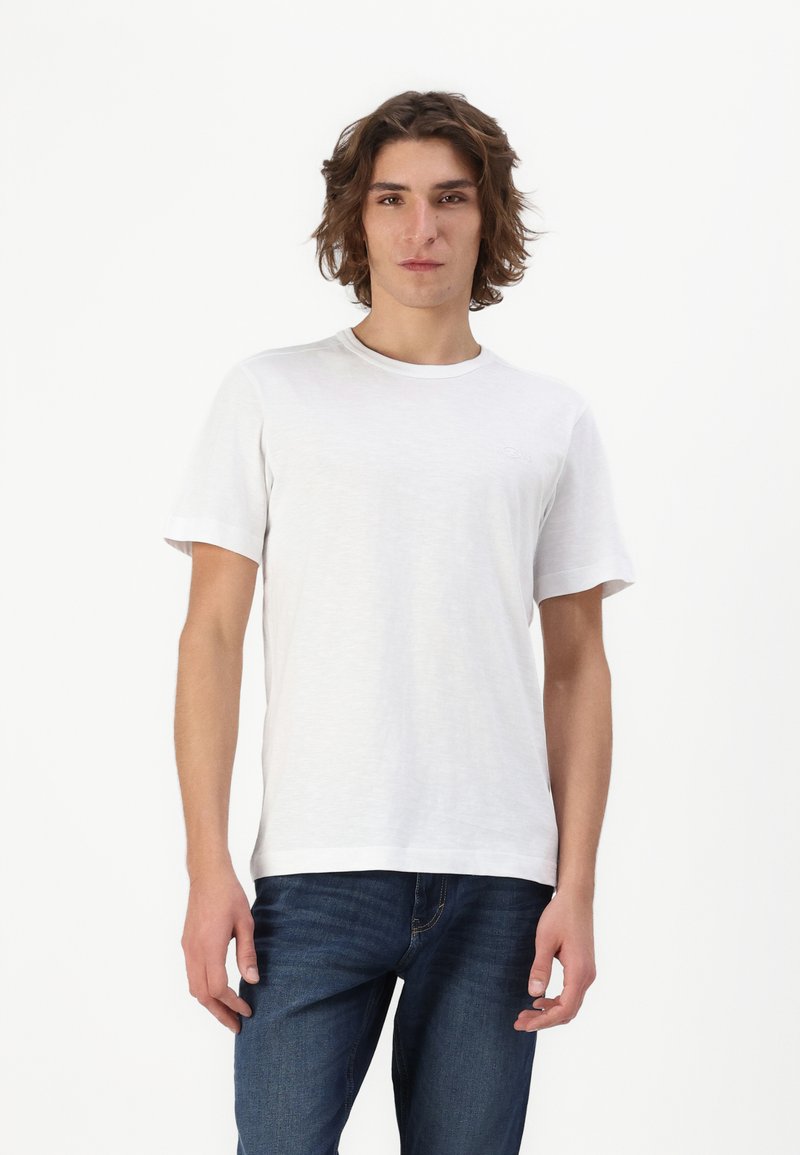 Hvid kortærmet T-shirt lavet af blødt, let materiale med rund halsudskæring. Modellen parrer den med blå jeans og står foran en hvid baggrund.