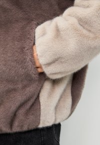 Giacca in pelliccia sintetica con pannelli a contrasto beige chiaro e marrone scuro. Presenta una texture morbida e tasche laterali.