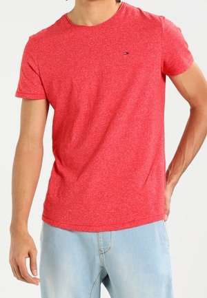 Mann trägt ein meliertes rotes kurzärmeliges T-Shirt und hellblaue Jeans, steht mit einer Hand hinter dem Rücken.