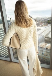 Blondidega naine, kes kannab kreemjat palmikutega kootud kampsunit ja valgeid pükse, hoides vastavat kampsunit ja beežikut kotti, seistes suure akna ääres.