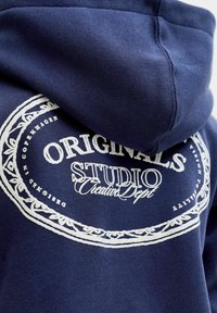 Námořnická modrá mikina s kapucí vyrobená z měkké látky, která má bílé kulaté logo s nápisy „ORIGINALS“, „STUDIO“ a dekorativním okrajem.