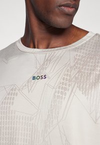Hellbeige T-Shirt mit geometrischem Muster, das dezente Texturen aufweist und ein mehrfarbiges "BOSS"-Logo auf der Brust hat.