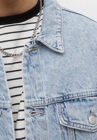 Gros plan sur une personne portant une veste en denim bleu clair par-dessus un t-shirt rayé noir et blanc avec un collier chaîne en argent.