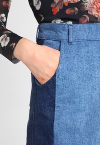 Jupe en denim de couleur bleue, avec une poche sur le côté gauche. Tissu texturé avec des teintes contrastantes dans un design à panneaux.