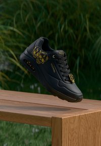 Μαύρο αθλητικό παπούτσι Skechers με χρυσά σχέδια κρανίου στο πλάι και κορδόνια, αιωρείται πάνω από ξύλινο παγκάκι με φόντο φυλλώματα.